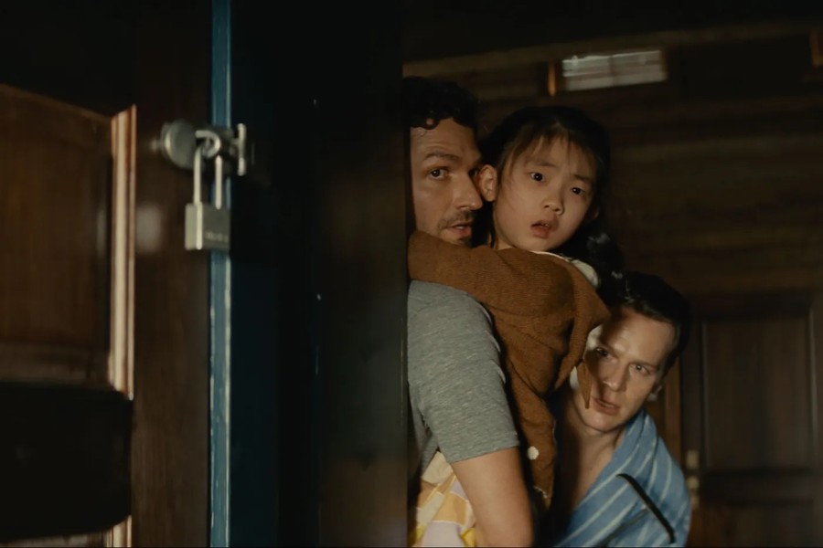 Actors Kristen Cui, Jonathan Groff and Ben Aldridge in the film “Knock at the Cabin.” They are hiding behind a corner in a cabin and looking anxiously to the right.