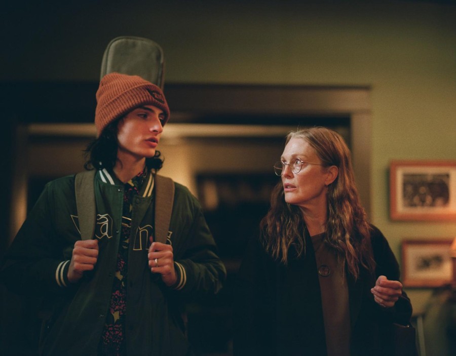 A teenager wearing a green jacket and a burnt orange beanie is standing, holding a guitar case on his back. A woman stands to the right of the teenager, wearing wire-frame glasses and black clothes. They are looking at one another.