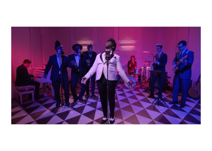 A singer wearing a white blazer in the middle of a parlor with black-and-white triangular tiles. There is a group of performers and a big band behind them.