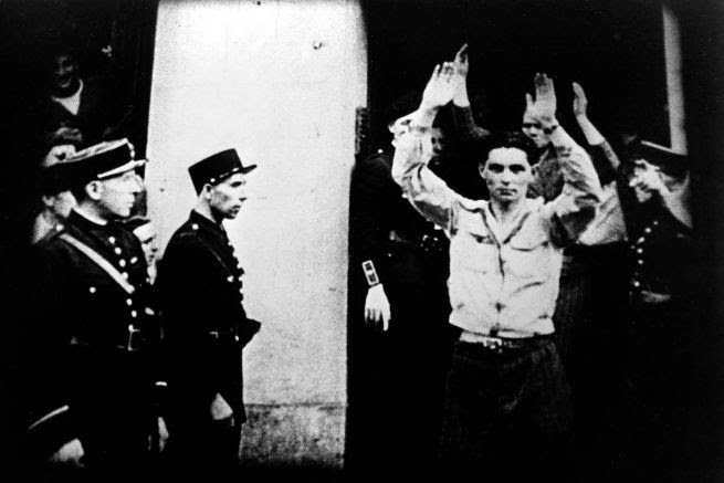A black and white photograph of people on the right side holding their hands up. They are surrounded by men in uniform.
