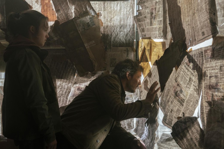 A man in a jacket peeks out between pieces of newspaper covering a window. A teenage girl stands behind the man and watches him as he looks out.