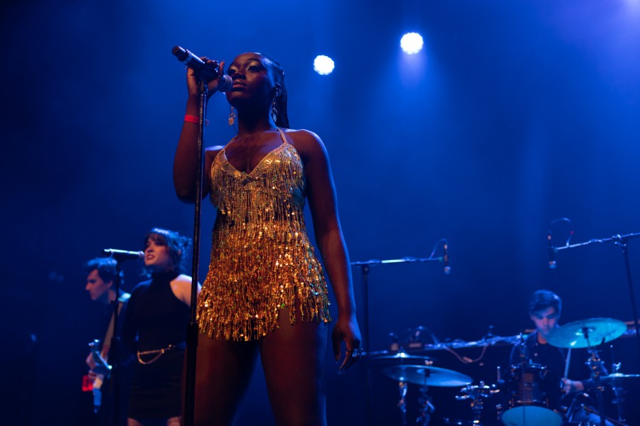 A woman wearing a gold dress stands beneath multiple blue spotlights, behind a microphone stand with one hand on the mic. Behind her is a backup singer dressed in black, a man behind a drum set and a man holding a guitar.