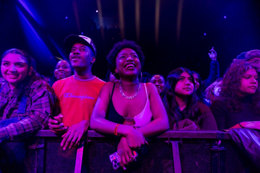 A line of people stand behind a metal barricade. They are standing under purple and blue spotlights, looking upward.