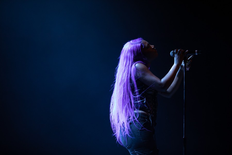 A woman with purple hair wearing black stands in front of a microphone stand, which she holds in one hand.