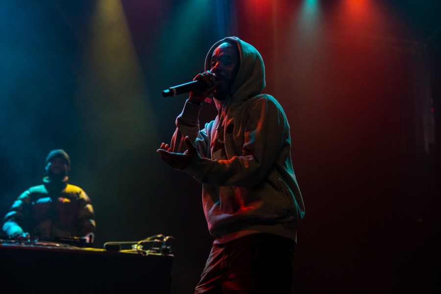 A man in a gray sweatshirt with the hood pulled up holds a microphone to his mouth under dim red, yellow and blue lights. Behind him is a man at a table with D.J. equipment.