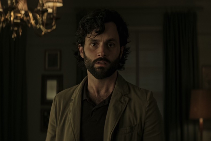 A man with long and curly hair and beard wearing a dark green jacket. He is in a dimly lit room.
