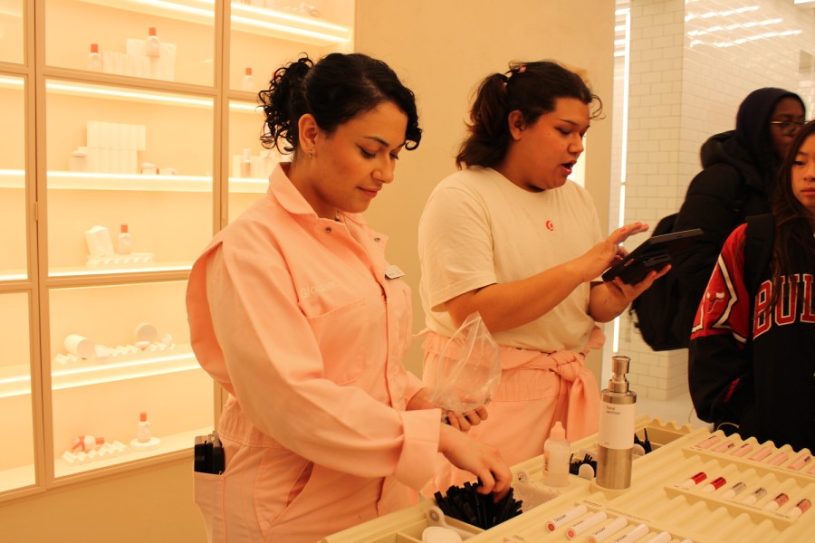 A photograph of the inside of Glossier SoHo, showing two store workers ringing up products for customers. The worker on the left wears a pastel pink jumpsuit and messy hair bun. The worker on the right wears a half unzipped pastel pink Glossier jumpsuit with a white Glossier t-shirt visible underneath.