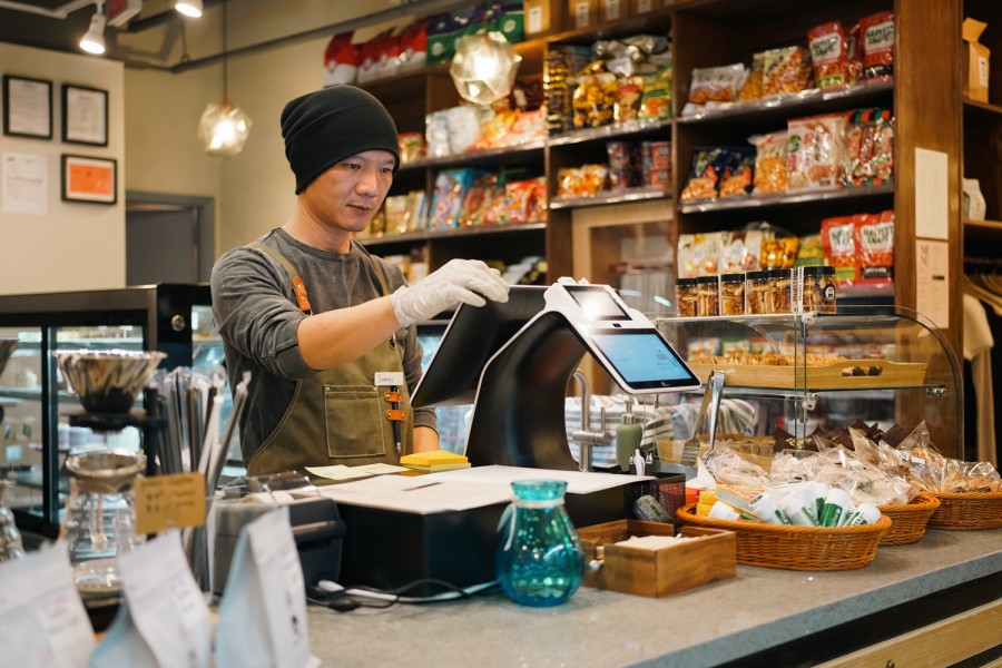A barista wearing an apron, a black beanie and white plastic gloves operates a cashier’s kiosk inside of a coffee shop. A shelf of snacks is in the background.