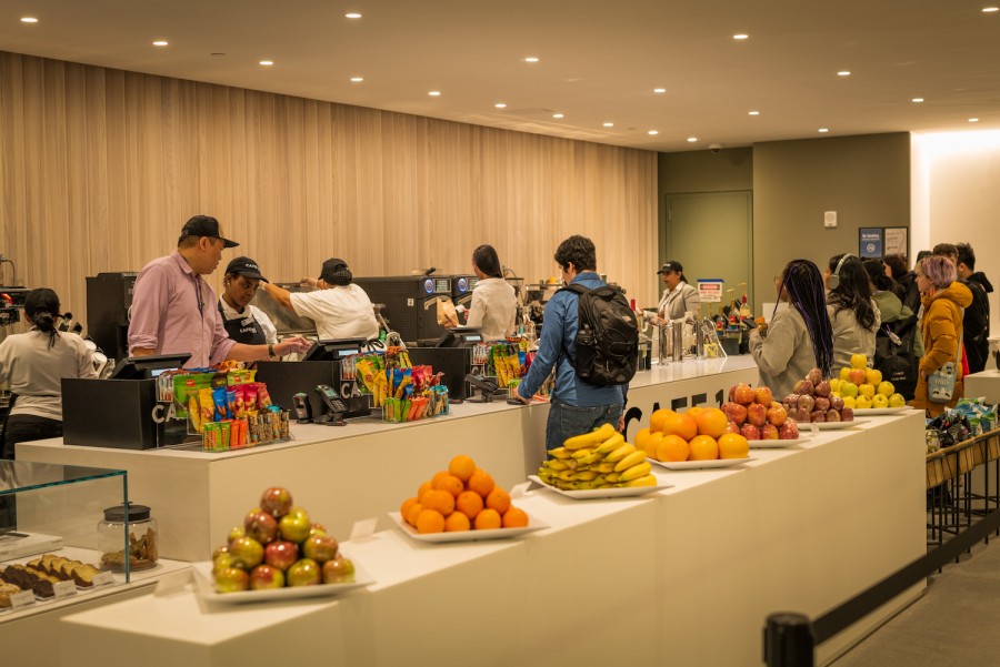 In the warmly-lit dining hall in the Paulson Center, a line of students wait next to the cashier counter. Seven plates of neatly-arranged fruits are placed on a white table.