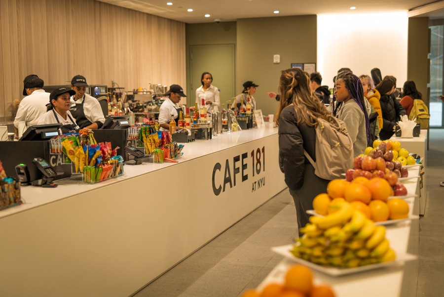 Students wait in a checkout line with baskets of fruit scattered along the dividing walls, and candy and snacks next to the cash registers with cashiers working behind.