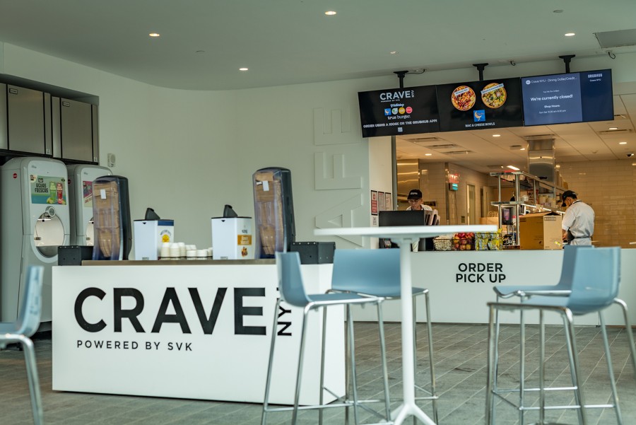 A set of high chairs and a table sit in front of a condiment bar bearing the words “Crave N.Y.U. Powered by S.V.K.” Behind the condiment bar is a counter reading “Order Pick Up” with two men dressed in white behind it working in a kitchen. Above the counter are three T.V.s displaying menu options and food photos.