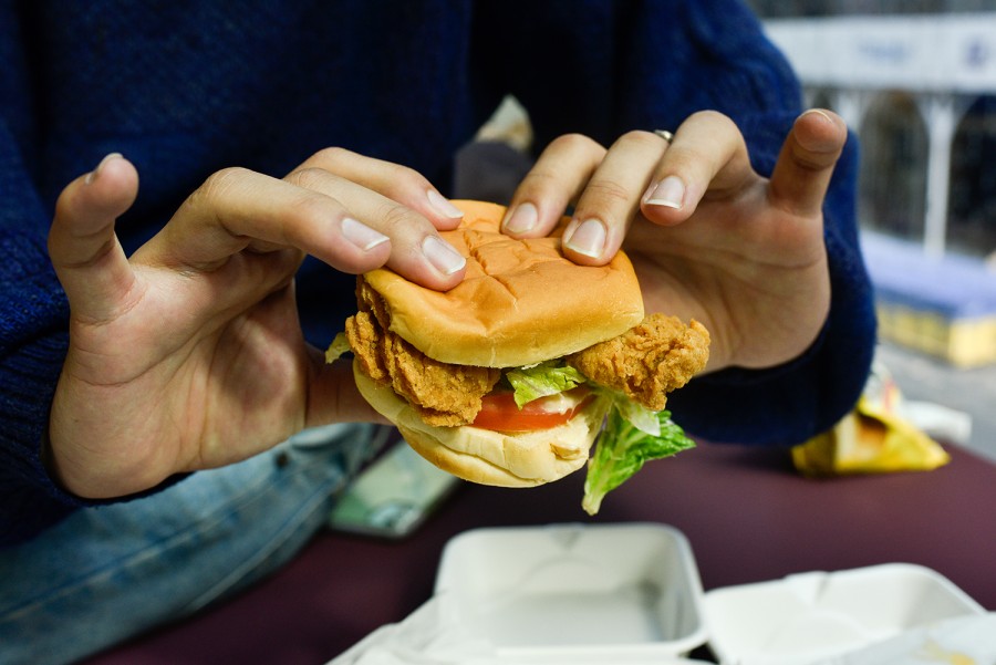 A person wearing blue holds a breaded chicken sandwich with tomato and lettuce on a bun in their hands. Below their hands are open white containers on a maroon table.