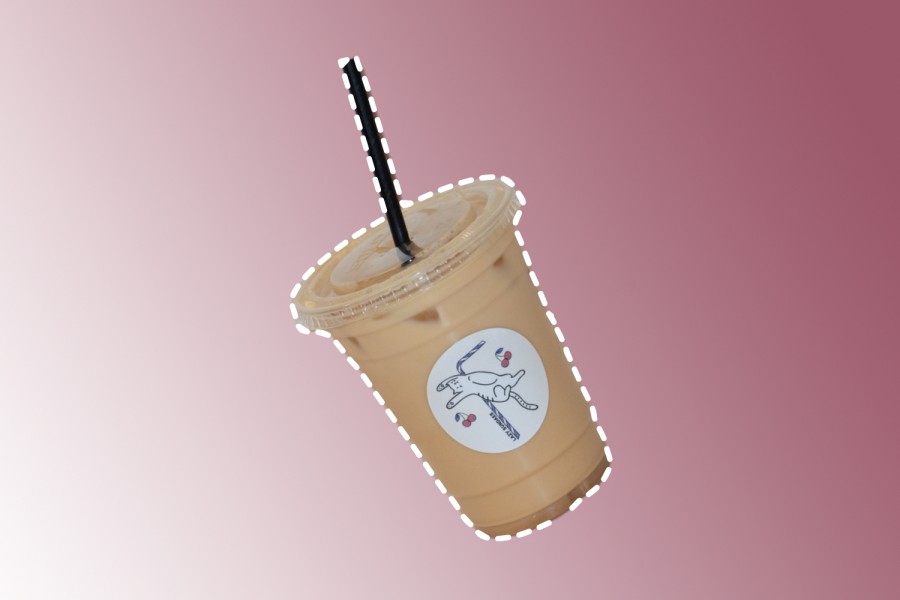 A tan plastic cup with a black straw against a pink gradient background. There is a white circular sticker on the middle of the cup. The design on the sticker is a purple and white striped straw with a white cat laying in between two pairs of cherries.