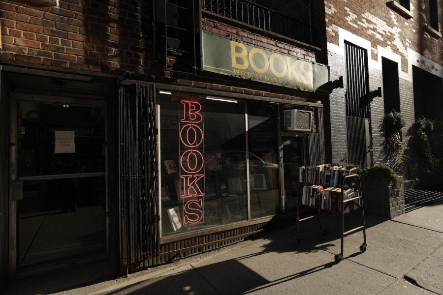 The storefront of Mercer Street Books and Records. Above the entrance is a green banner with yellow text that reads “Books. Mercer Street Books. 206 Mercer.” Behind the storefront window is a red neon sign that reads “Books.” A shelf full of books is on the sidewalk of Mercer Street.