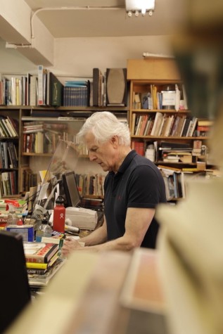 With an indistinct stack of books in the foreground, an elderly man stands behind the checkout counter of Mercer Street Books and Records.