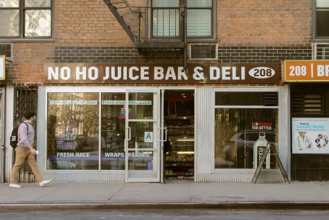 The storefront of NoHo Juice Bar and Deli. A pedestrian walks by the storefront, looking through the window into the store. On the window is text that reads “Fresh Juice. Wraps. Paninis.”