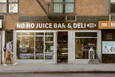 Shop window of NoHo Juice Bar and Deli.  A passer-by walks past a shop window, peering through a shop window.  There is text on the window that reads: “Fresh juice.  Wraps.  Panini.