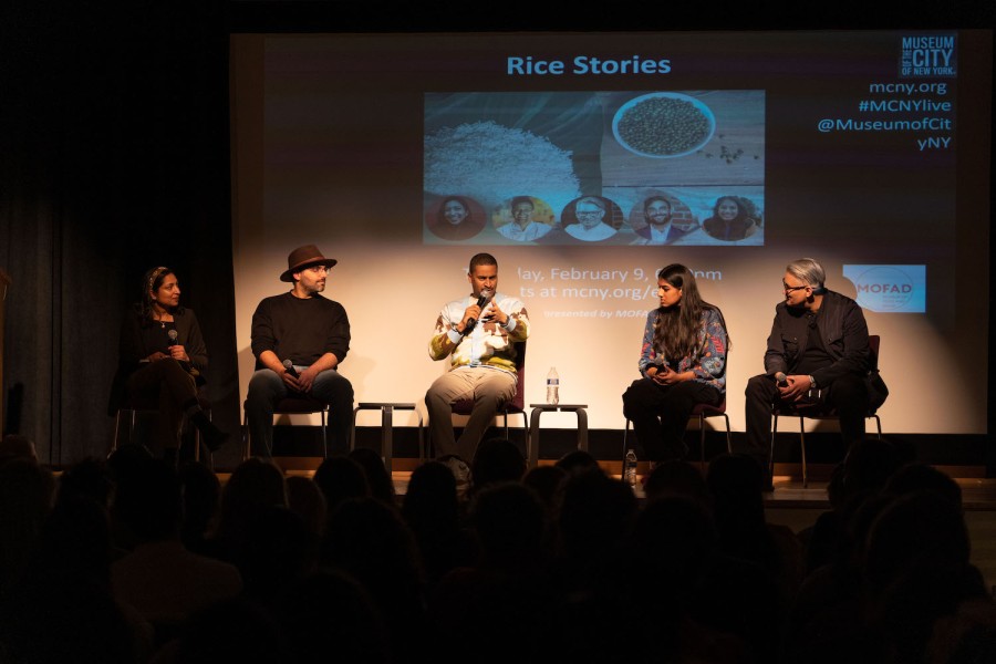 Five panelists speaking on a stage in dim lighting. There is a projector screen behind them with the text “Rice Stories” that indicates the event is held at the Museum of the City of New York.