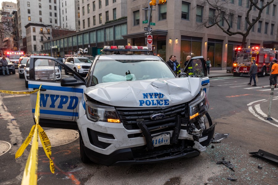A police car sits in the middle of the intersection between east thirteenth street and university place with its driver’s side airbags blown, and yellow police tape reads “Do Not Cross,” draped on its left side. The front of the vehicle is severely damaged, with shattered glass and parts of the car in pieces on the pavement. Behind it are civilians and their vehicles along with other police cars and a firetruck.