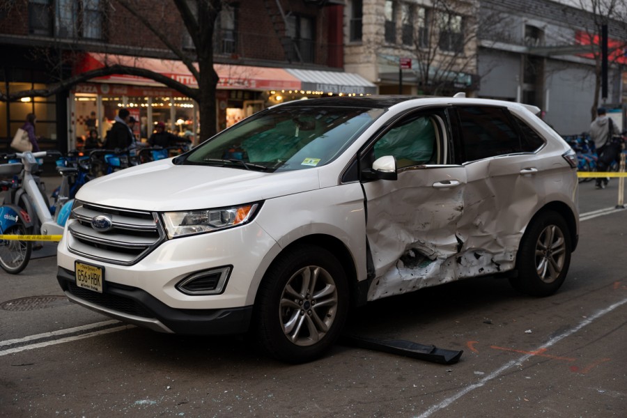 A white car is parked in the middle of the street in front of a row of Citi Bikes.  The driver's and left passenger's doors were severely damaged, and the driver's airbag deployed.