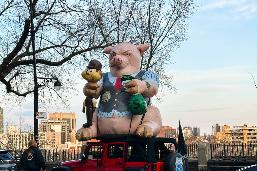 A giant inflatable pig display that is wearing a blue short-sleeved shirt and gray vest with a pocket full of money sits on a red Jeep, holding a squeezed labor worker character in one hand and a green bag with money sign in the other hand.