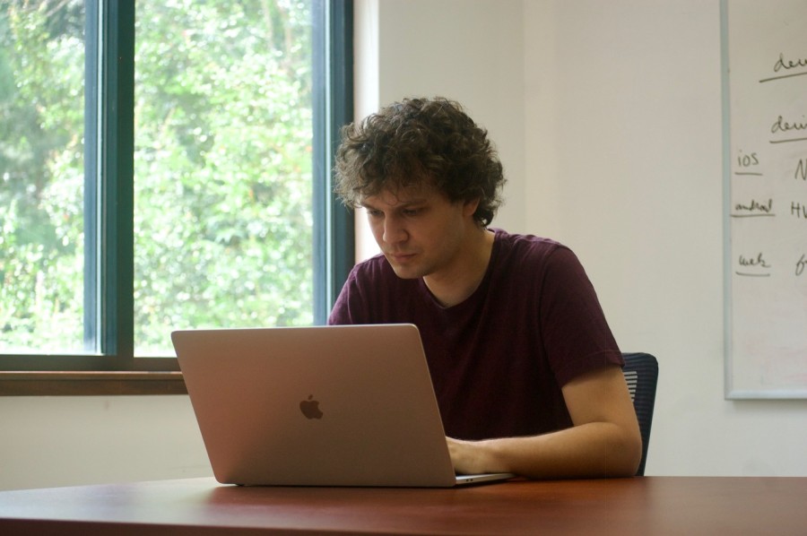 Joe Puccio, the co-founder of Coursicle, wearing a maroon shirt, sits behind a MacBook Pro on a table. Behind him is a window with sunlight streaming in.