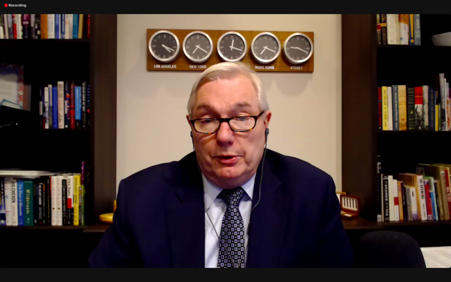 A man wearing a dotted tie, a white shirt and a black suit jacket speaks in an online conference session. Behind him are a bookshelf and five clocks displaying times in different time zones.