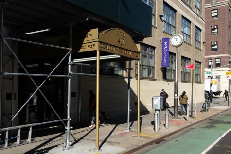 The entrance to N.Y.U’s College of Dentistry under scaffolding. A sign for twenty-fifth street is seen to the right of the building, and a clock is mounted on a pole on the sidewalk.