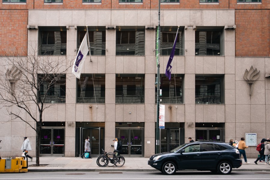 The outside of N.Y.U.s Palladium residence hall, with two N.Y.U. flags and two N.Y.U. torches on the side of the building. A car is parked in front and people walk past.