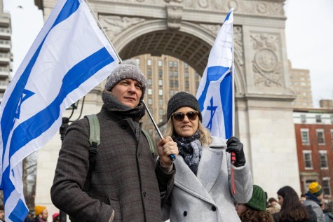 A man in a dark gray jacket and a woman in a light gray jacket, both wearing gray winter hats, hold full-size Israeli flags on poles. They are standing in front of the Washington Square Arch in Washington Square Park.