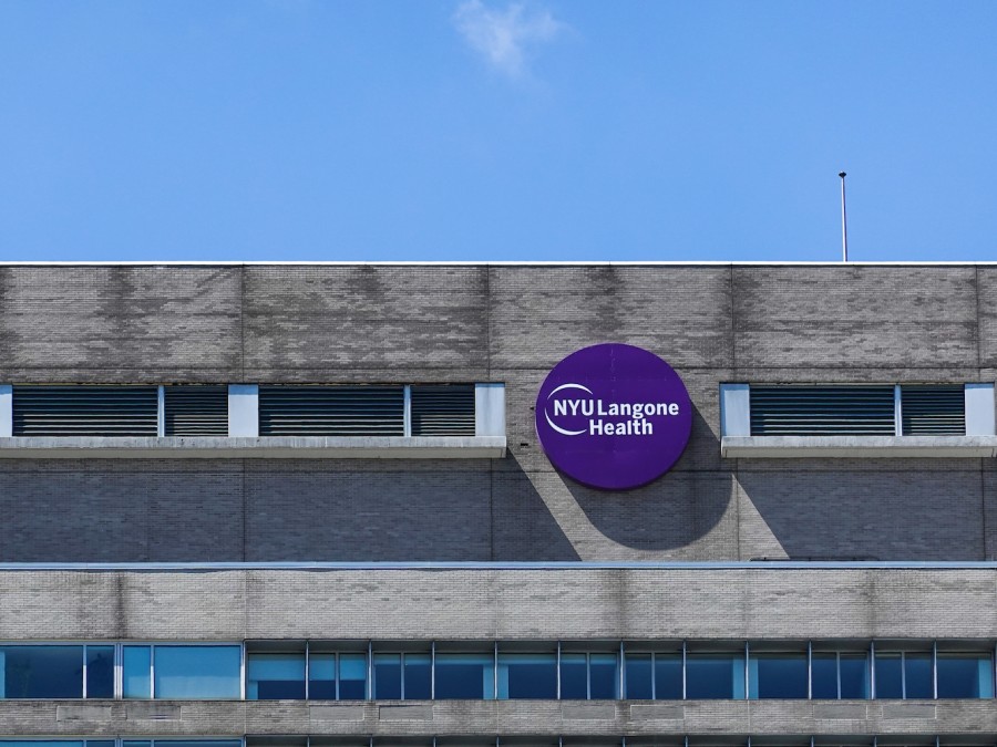 The exterior of the top floor of a building with a circular, purple N.Y.U. Langone Health sign. The blue sky is seen above.