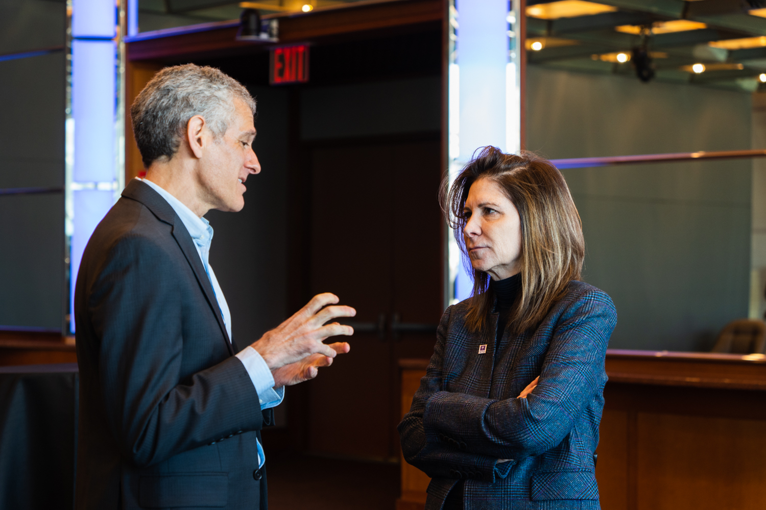 N.Y.U president-designate Linda Mills in a conversation with Cecil Scheib, the university’s chief sustainability officer, who is wearing a black suit. Mills is wearing a gray jacket with an N.Y.U pin on the collar.