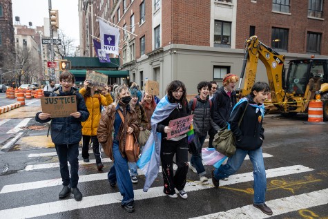 Tens of high school students march across a sidewalk in front of a building with a NYU banner on an overcast day. Two people are draped in the transgender flag, wearing it like a cape