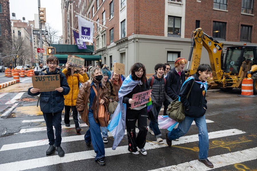 Tens+of+high+school+students+march+across+a+sidewalk+in+front+of+a+building+with+a+NYU+banner+on+an+overcast+day.+Two+people+are+draped+in+the+transgender+flag%2C+wearing+it+like+a+cape