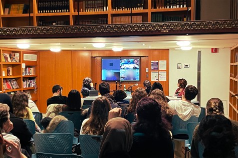 In a room filled with bookshelves, a crowd of attendees sits in front of a T.V. screen showing four panels of speakers.