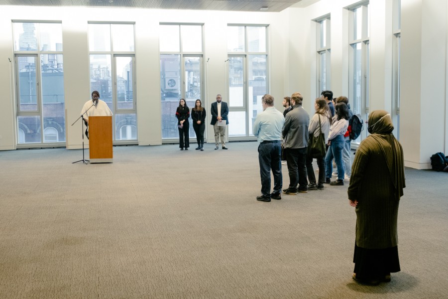A speaker delivers a speech from behind a podium. Observers stand in a semicircle in front of the speaker. Vertical floor-to-ceiling windows are behind the speaker.