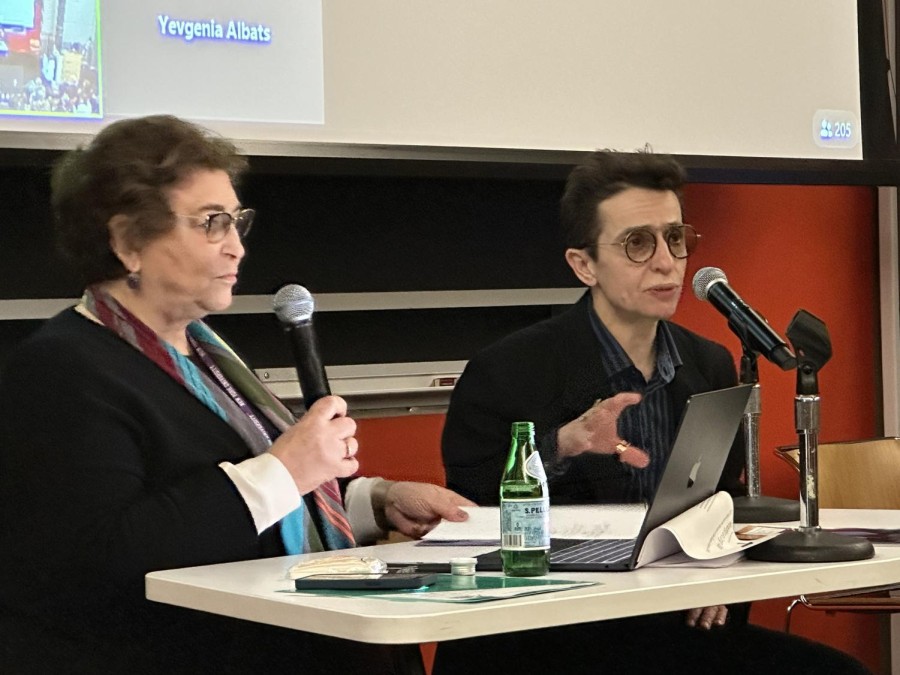 Two people sit behind a white table with a projector screen behind them. On the left is Yevgenia Albats, wearing a black jacket and a colorful scarf while holding a microphone. On the right is Masha Gessen, wearing a black jacket with a microphone in front of them.