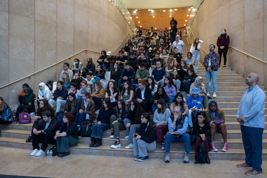 A crowd of people sit on a marble staircase. Many of them have their eyes closed and their heads facing down to observe a moment of silence. Many of them are looking to the left, toward a podium, which is out of frame.