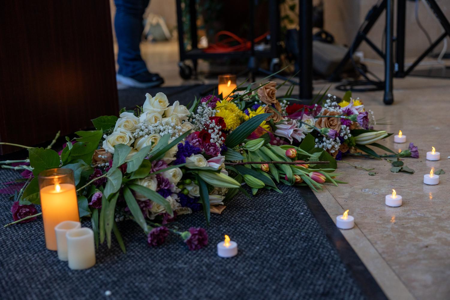 Various bouquets of flowers placed on a marble floor and on top of a grey carpet, in front of a podium. Around the flowers are battery-powered candles and two tall lit candles.