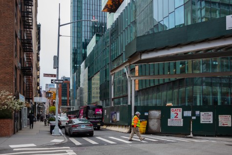 A tall building complex with protruding glass windows under construction. Across the street from the construction site is a row of buildings with brown brick exteriors. The road sign indicates that the building is on Bleecker Street.