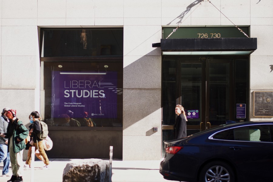 This photo shows the brightly-lit exterior of N.Y.U’s Liberal Studies building. In the bottom right of the image, a pedestrian is walking by the building entrance, and a black car is parked in the foreground of the image. The shadows of the entrance are shown on the light beige walls, and a purple sign with white text that reads “Liberal Studies” is hung inside a big square window on the left of the image.