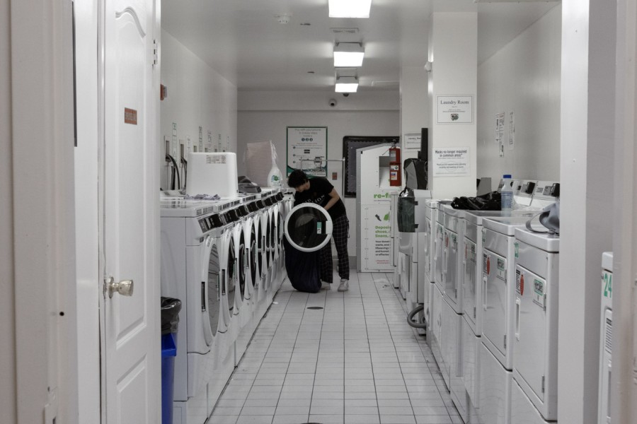 A+student+dressed+in+a+black+t-shirt+and+black+checkered+sweatpants+does+laundry+in+the+Coral+Tower+residence+hall+laundry+room.+The+laundry+room+consists+of+two+rows+of+white+washers+and+dryers.