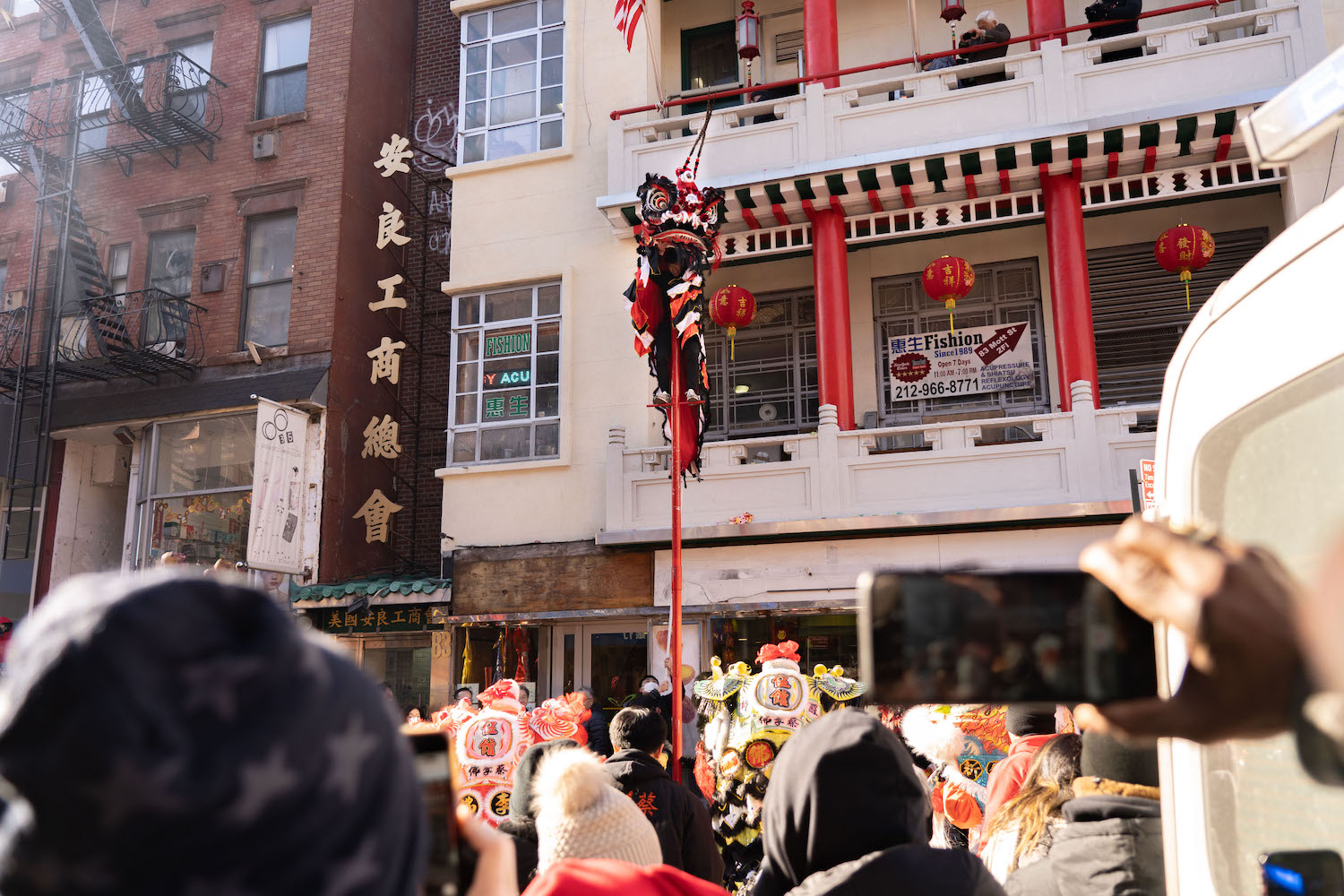 A lion dancer standing on top of a two-story tall pole in front of a crowd on the street.