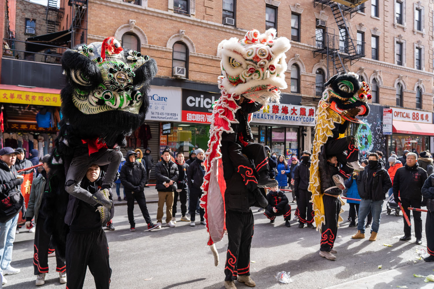 Three lion dance performers stand side by side among a crowd. Each lion is wielded by two people, one standing on the shoulder of another.
