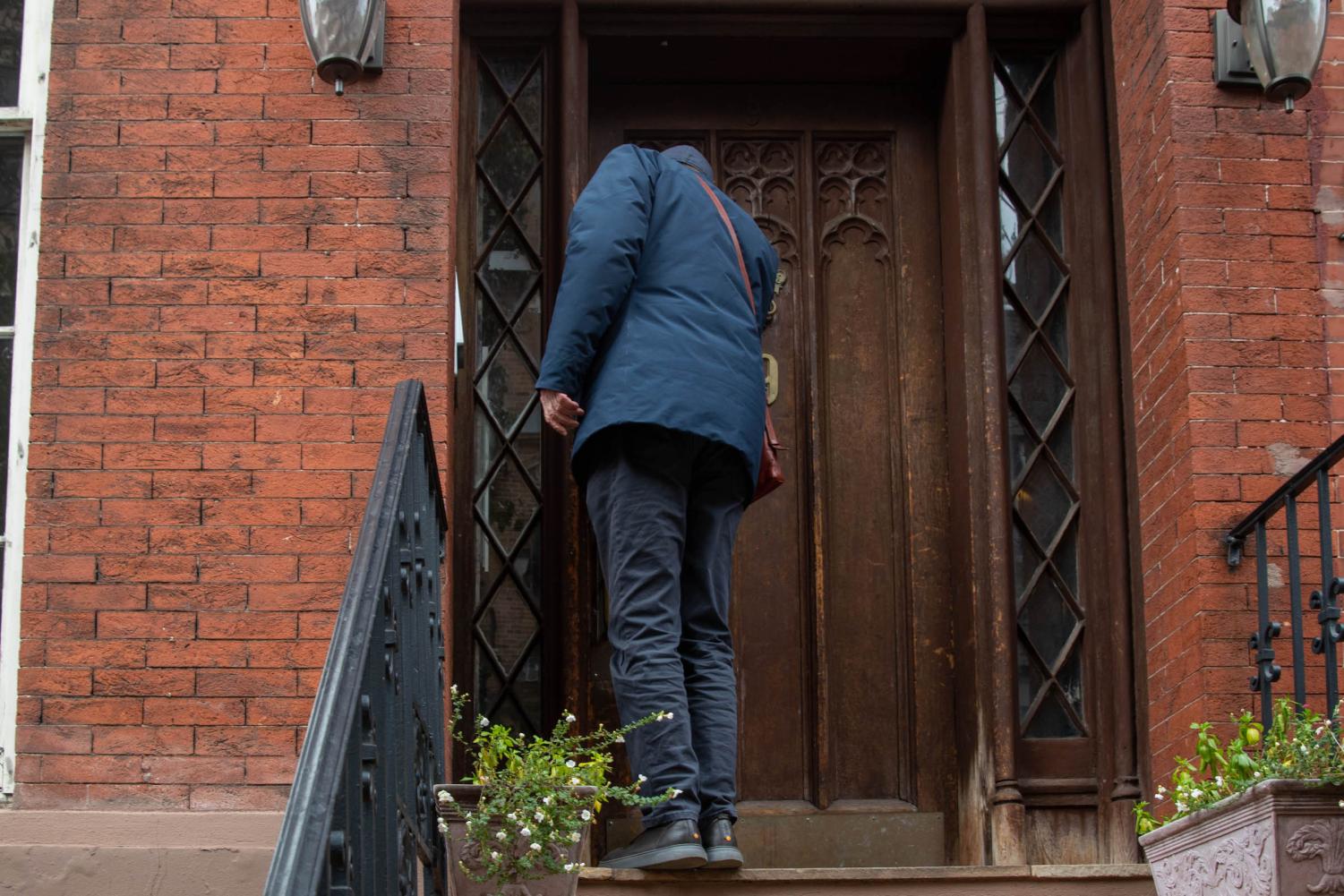 Alan Grossman opens the door to his brownstone apartment while wearing a brown satchel and a navy blue jacket. There are potted flowers on the stairs leading to the door.
