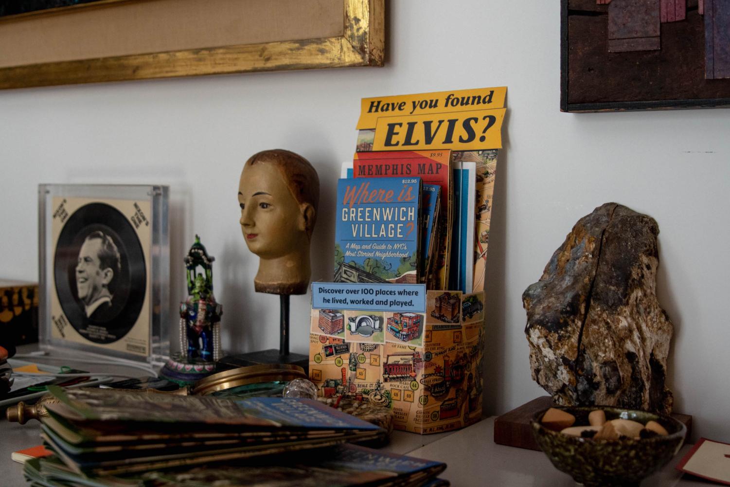 A close-up shot of a group of decorations in Grossman's studio atop a table, framed against a white wall. It includes photographs, maps, framed artwork and figurines.
