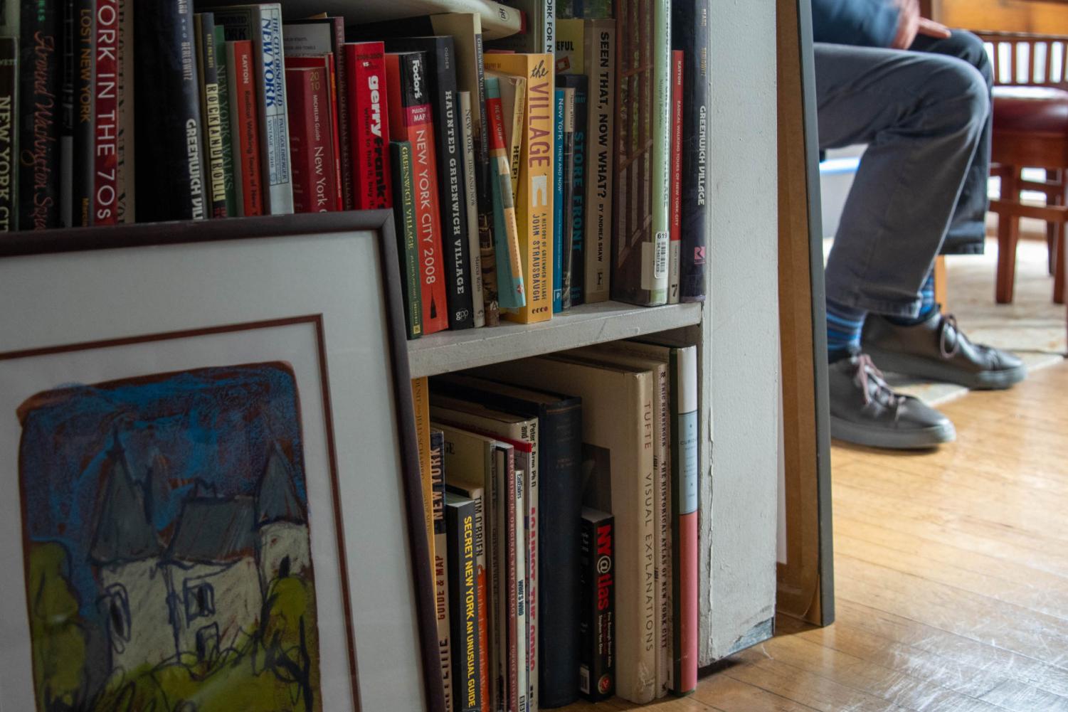 The bottom of Alan Grossman's bookshelf is full of colorful books about New York, and there is a framed painting leaning against it. In the background, Grossman sits at a table wearing navy blue pants and shoes.