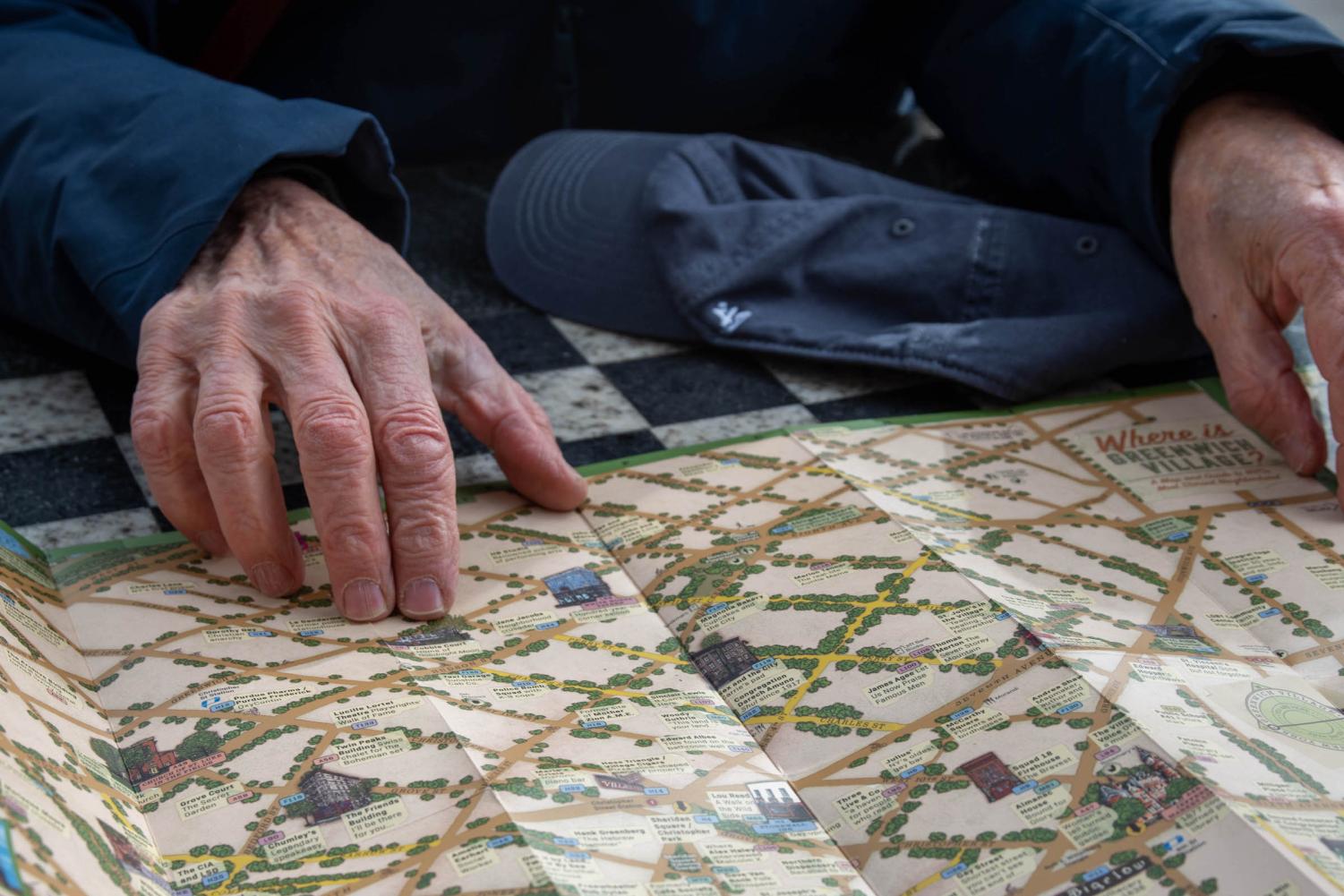 Alan Grossman's hands are shown placed on his map of Greenwich Village, on top of a chess table. He is wearing a navy blue jacket, and his navy blue baseball cap is on the table.