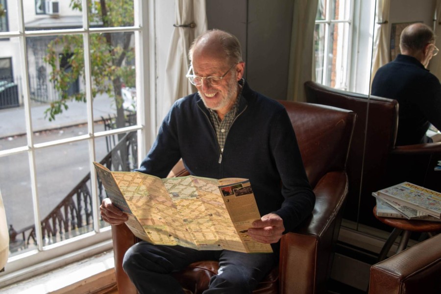 Alan Grossman is pictured, wearing jeans, a navy blue quarter-zip sweater, a blue-and-white button-up shirt and clear glasses. He is sitting on a brown leather couch and holding a map. Behind him is a window with a white curtain facing the street.