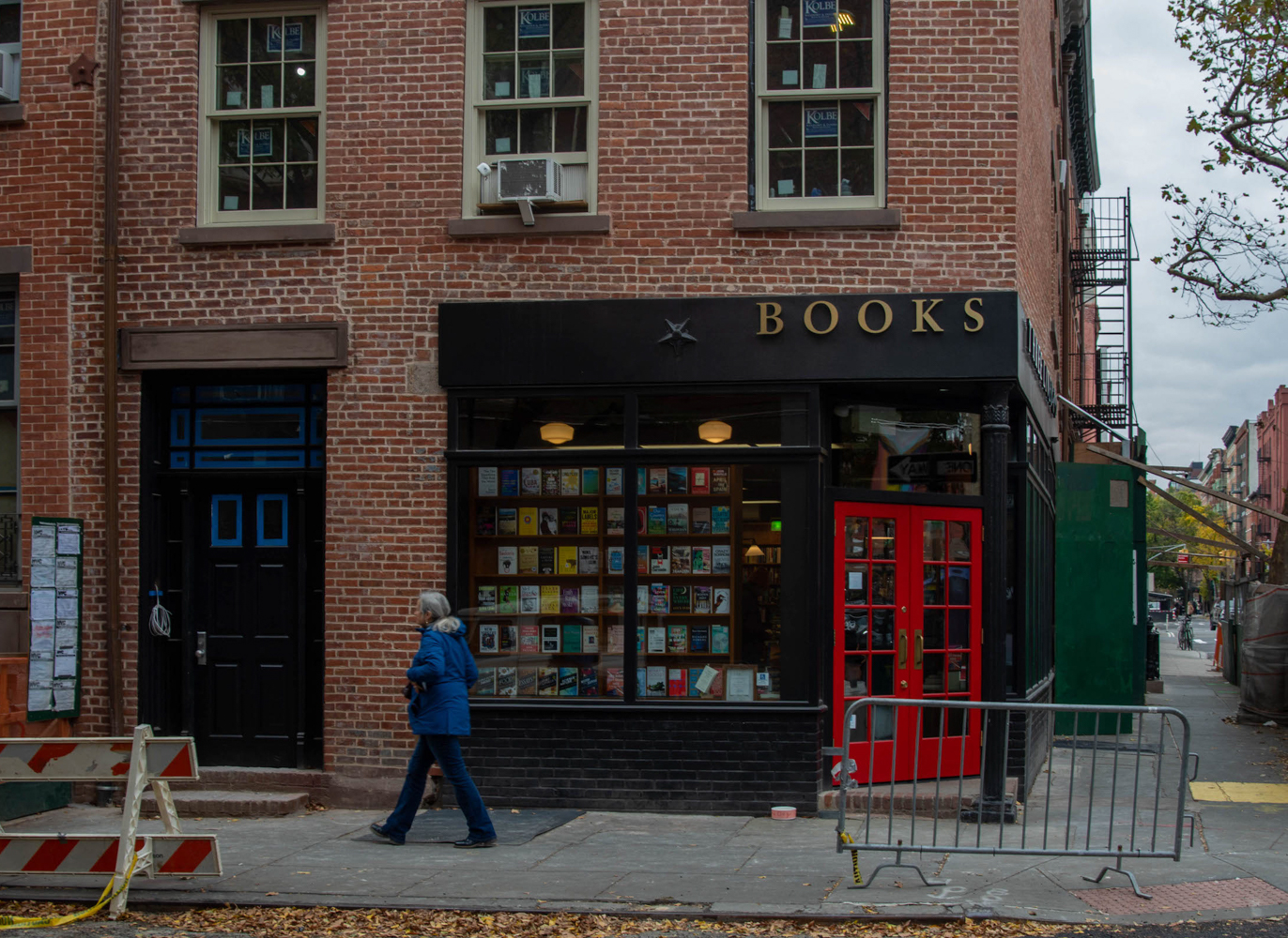 A person with long, gray hair is wearing blue jeans and a blue jacket while walking down the sidewalk. Behind them is the corner of Third Life Books with a bright red door and a window display of books. Construction equipment is on the sidewalk and street around them.